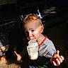 baby, blue_eyes, bottle, brown_hair, child, colorful_clothing, dramatic_lighting, feeding, gray_shirt, indoors, milk, orange_trim, patterned_pants, portrait, shelving, shoes, sitting, sunlight, toddler, wood_floor