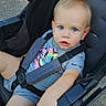 baby, blond_hair, blue_eyes, child, colorful_graphic, face, family, gray_shirt, looking_at_camera, outdoor, pavement, playful, portrait, safety_straps, seat_harness, sitting, stroller, sunlight, toddler, travel