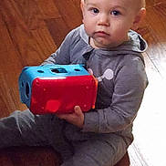 Hayes is registered to the contest to win money with this photo: baby, child, toddler, sitting, toy, colorful_toy, red, blue, gray_clothing, wooden_floor, indoors, portrait, face, eyes, serious, looking_at_camera, bald, infant, playtime, home