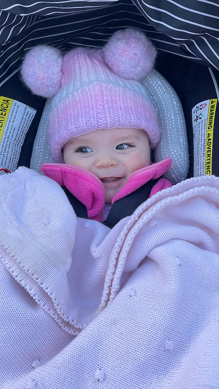 Kylie is registered to the contest to win money with this photo: baby, baby_products, baby_toddler_clothing, beanie, cap, car_seat, cheek, comfort, electric_blue, headwear, knit_cap, magenta, person, purple, sitting, sleeve, smile, toddler, violet, winter