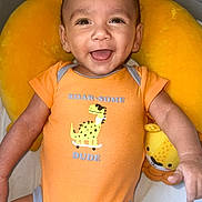 Giovanni is registered to the contest to win money with this photo: baby, child, smiling, onesie, orange_clothing, dinosaur_graphic, stuffed_toy, yellow_cushion, happy, infant, cute, portrait, indoor, baby_boy, playful, lying_down, closeup, face, expression, soft_toy