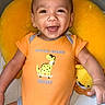baby, child, smiling, onesie, orange_clothing, dinosaur_graphic, stuffed_toy, yellow_cushion, happy, infant, cute, portrait, indoor, baby_boy, playful, lying_down, closeup, face, expression, soft_toy