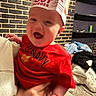 baby, child, red_shirt, old_navy, hat, paper_hat, in_n_out_burger, smiling, happy, indoor, living_room, brick_wall, person, hand, holding, furniture, couch, clothing, casual, portrait