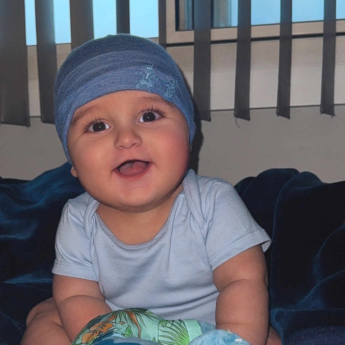 Matthias -Alexander joined the competition — help win amazing prizes! baby, bed, blanket, blue_beanie, child, clothing, cute, happy, home, indoor, infant, person, plush_toy, portrait, seated, smiling, soft_toy, vertical_blinds, window, young_child