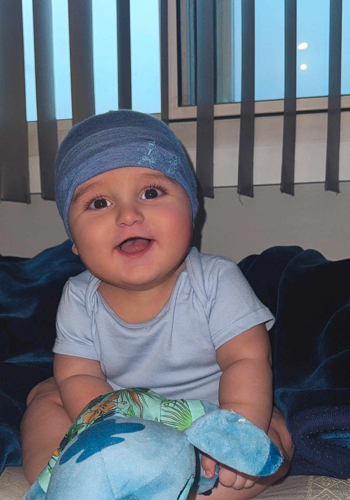 Matthias -Alexander joined the competition — help win amazing prizes! baby, infant, blue_beanie, smiling, plush_toy, bed, blanket, window, vertical_blinds, indoor, child, cute, happy, portrait, person, clothing, soft_toy, seated, home, young_child