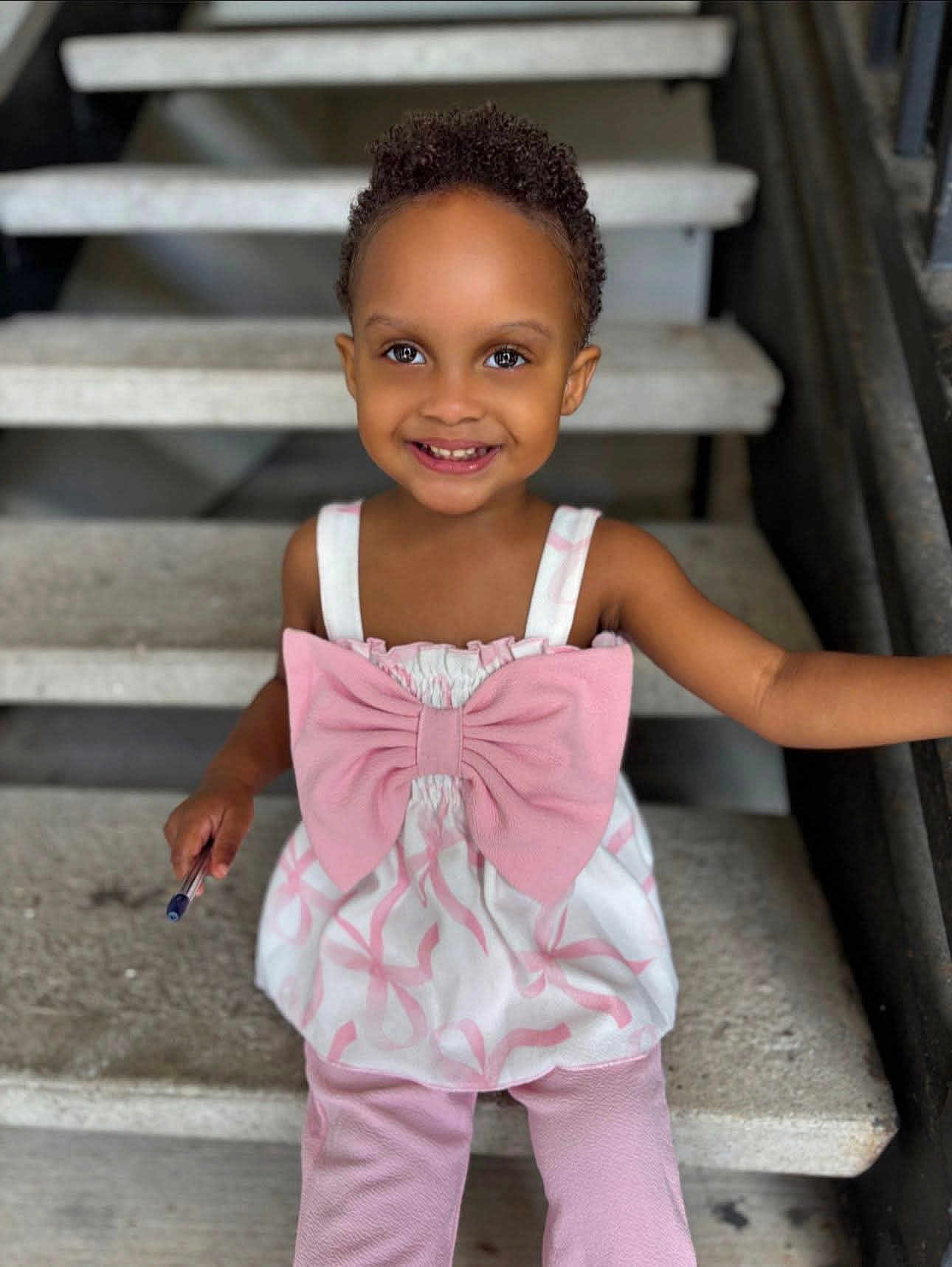Viviana Rose joined the competition — help win amazing prizes! child, toddler, smile, face, pink_clothing, bow, pen, stairs, concrete, sitting, happy, person, portrait, short_hair, indoors, railing, cute, bright_eyes, casual_clothing, young_child