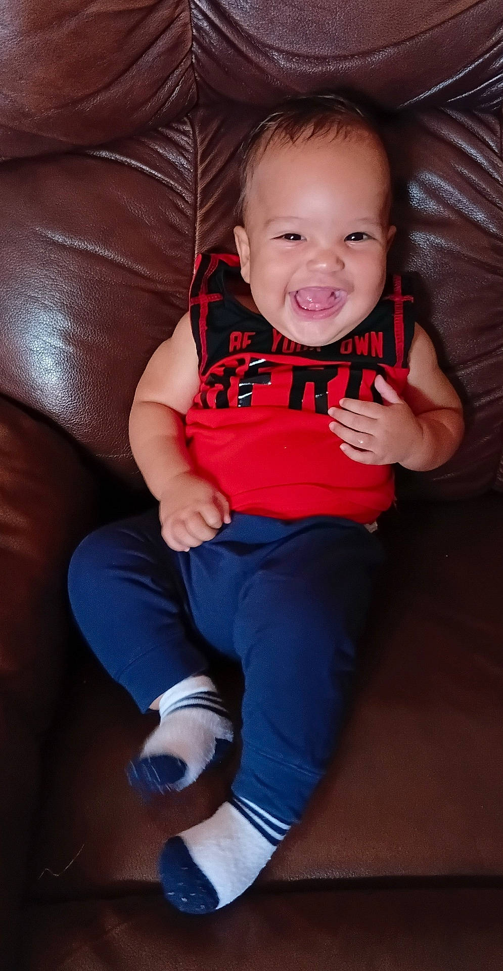 Angel is registered to the contest to win money with this photo: baby, black_clothing, blue_pants, child, close_up, crossed_legs, face, furniture, happy, home, indoors, joyful, laughing, leather_couch, portrait, red_shirt, sitting, smiling, socks, toddler