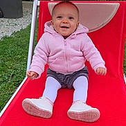 Louise a rejoint le concours — aidez-le/la à gagner de superbes lots ! baby, child, pink_hoodie, white_tights, shoes, red_lounge_chair, outdoor, grass, smiling, happy, sitting, person, face, footwear, garden, casual_clothing, cute, infant, playful, daylight