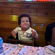 Julius joined the competition — help win amazing prizes! toddler, child, high_chair, restaurant, bread, food, smile, happy, wood, table, neon_sign, cozy, indoor, person, clothing, curly_hair, plate, blue_bottle, wall, casual