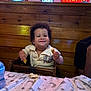 toddler, child, high_chair, restaurant, bread, food, smile, happy, wood, table, neon_sign, cozy, indoor, person, clothing, curly_hair, plate, blue_bottle, wall, casual