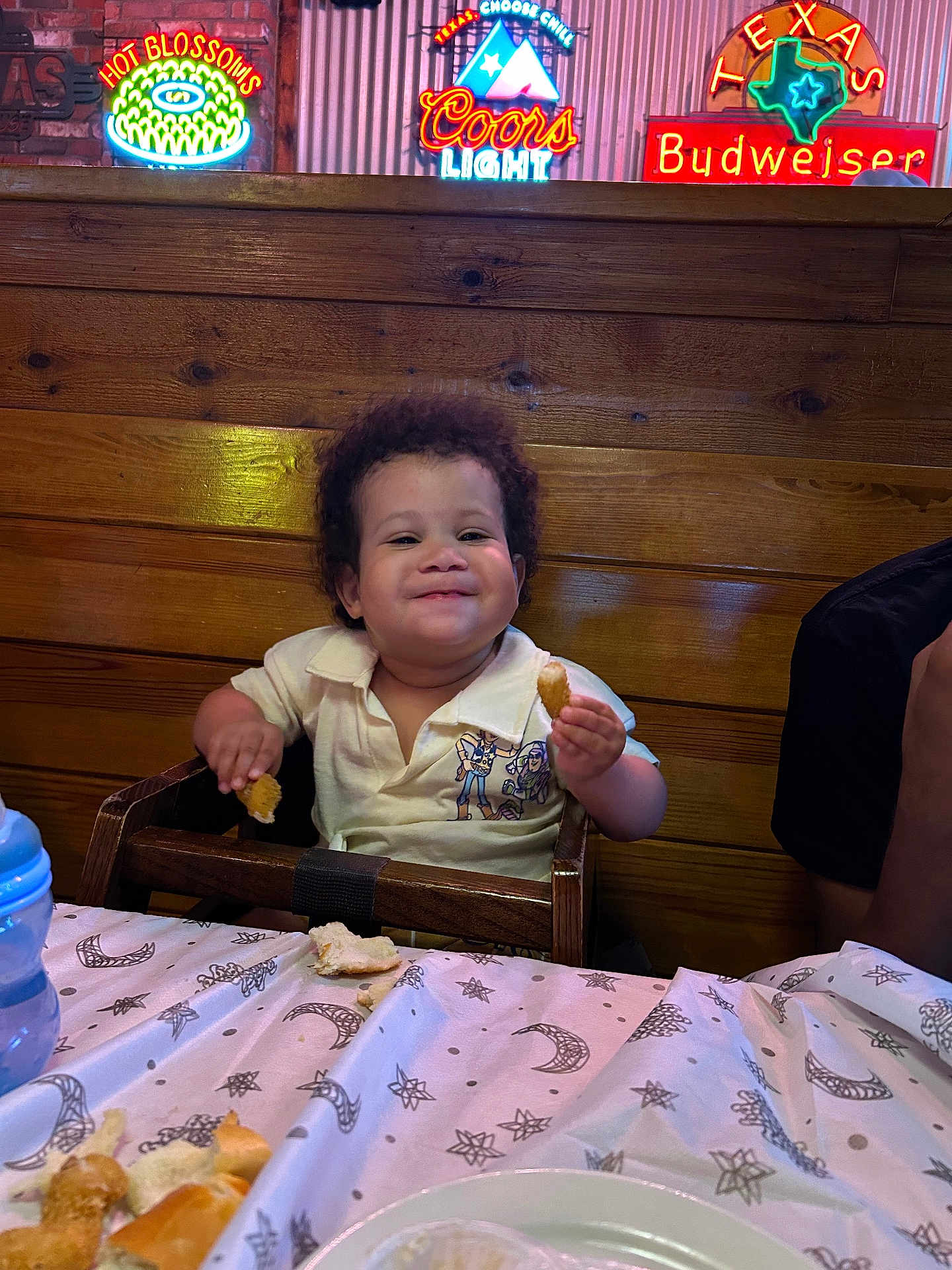 Julius joined the competition — help win amazing prizes! toddler, child, high_chair, restaurant, bread, food, smile, happy, wood, table, neon_sign, cozy, indoor, person, clothing, curly_hair, plate, blue_bottle, wall, casual