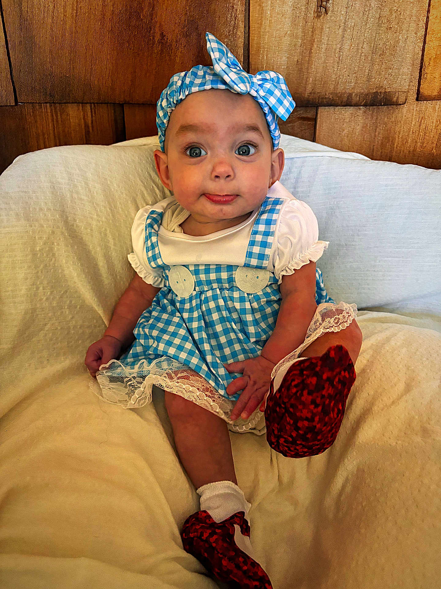 Lola is registered to the contest to win money with this photo: baby, child, blue_dress, headband, red_shoes, sitting, cushion, white_socks, wooden_background, indoor, cute, portrait, infant, clothing, fabric, face, expression, small_hands, furniture, person