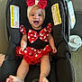 baby, child, car_seat, smiling, red_bow, polka_dot, headband, black_clothing, red_skirt, happy, indoors, warning_label, footwear_free, seatbelt, cute, infant, person, portrait, sitting, toy_book