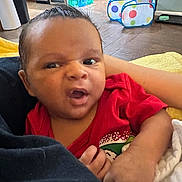 Bryson is registered to the contest to win money with this photo: baby, infant, child, face, expressive_face, onesie, red_clothing, hand, arm, blanket, towel, indoor, kitchen, refrigerator, floor, play_tent, polka_dot, sitting, portrait, cozy