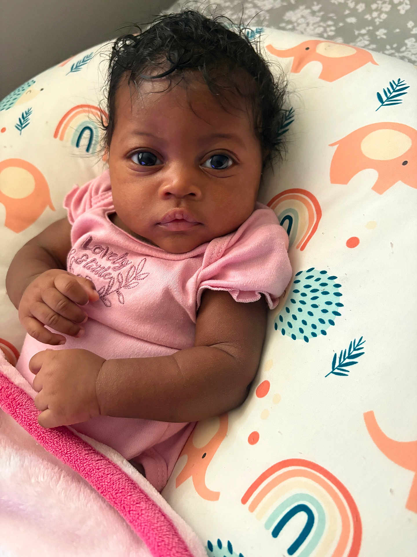 Clover is registered to the contest to win money with this photo: baby, infant, child, pink_clothing, cushion, rainbow_pattern, elephant_pattern, blanket, curly_hair, closeup, portrait, indoor, cute, small_hands, soft_texture, resting, looking_at_camera, adorable, peaceful, young