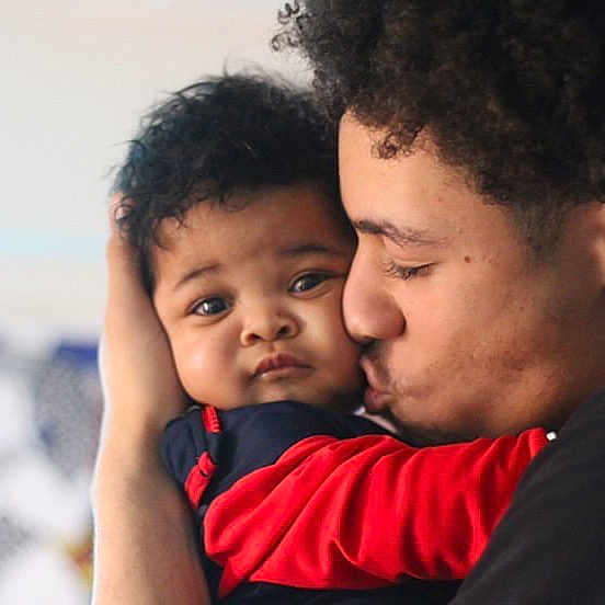 Cairo is registered to the contest to win money with this photo: baby, child, infant, person, face, love, affection, family, cuddle, kiss, curly_hair, red_clothing, black_clothing, portrait, close_up, indoor, skin, young_adult, expression, bond