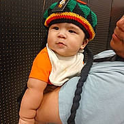 Ezekiel is registered to the contest to win money with this photo: baby, child, person, hat, knit_hat, orange_shirt, bib, braids, arm, adult, holding, curious, indoor, portrait, face, cute, chubby, black_hair, closeup, background