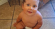 Frankie is registered to the contest to win money with this photo: baby, child, curly_hair, smile, diaper, floor, tile, kitchen, cabinet, happy, cute, sitting, indoors, person, skin, infant, young_child, one_person, looking_up, joyful