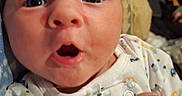 Rian is registered to the contest to win money with this photo: baby, child, close_up, clothing, cute, expression, face, furniture, hand, head, indoor, infant, newborn, onesie, open_mouth, person, portrait, skin, wide_eyes, young
