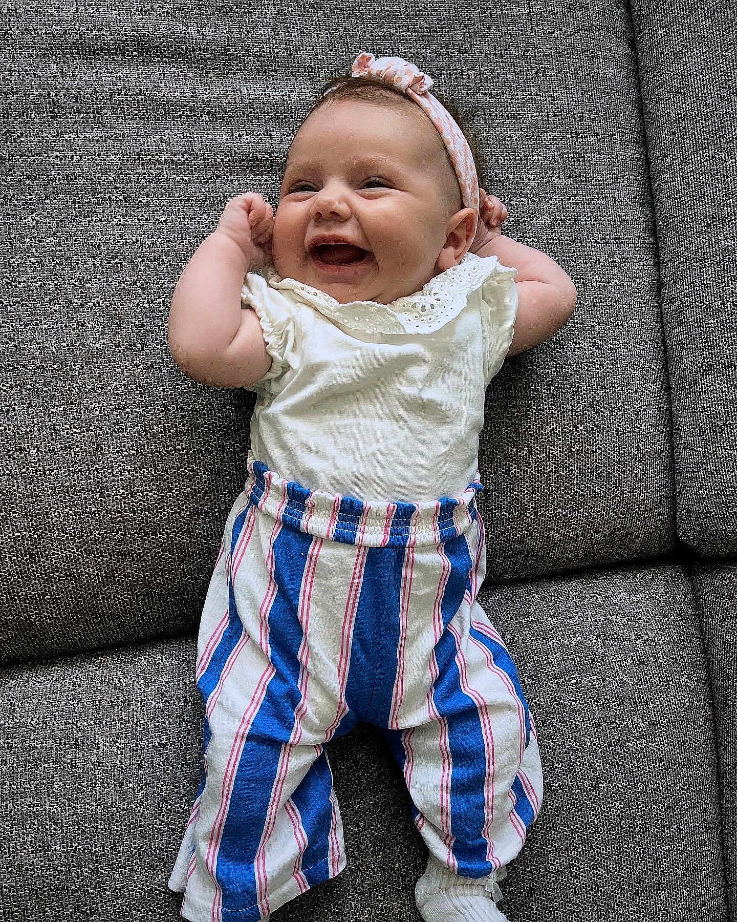 Victoire a rejoint le concours — aidez-le/la à gagner de superbes lots ! baby, smiling, couch, gray, striped_pants, headband, white_top, infant, happy, indoor, person, clothing, fabric, cute, playful, child, portrait, sitting, toddler, relaxing