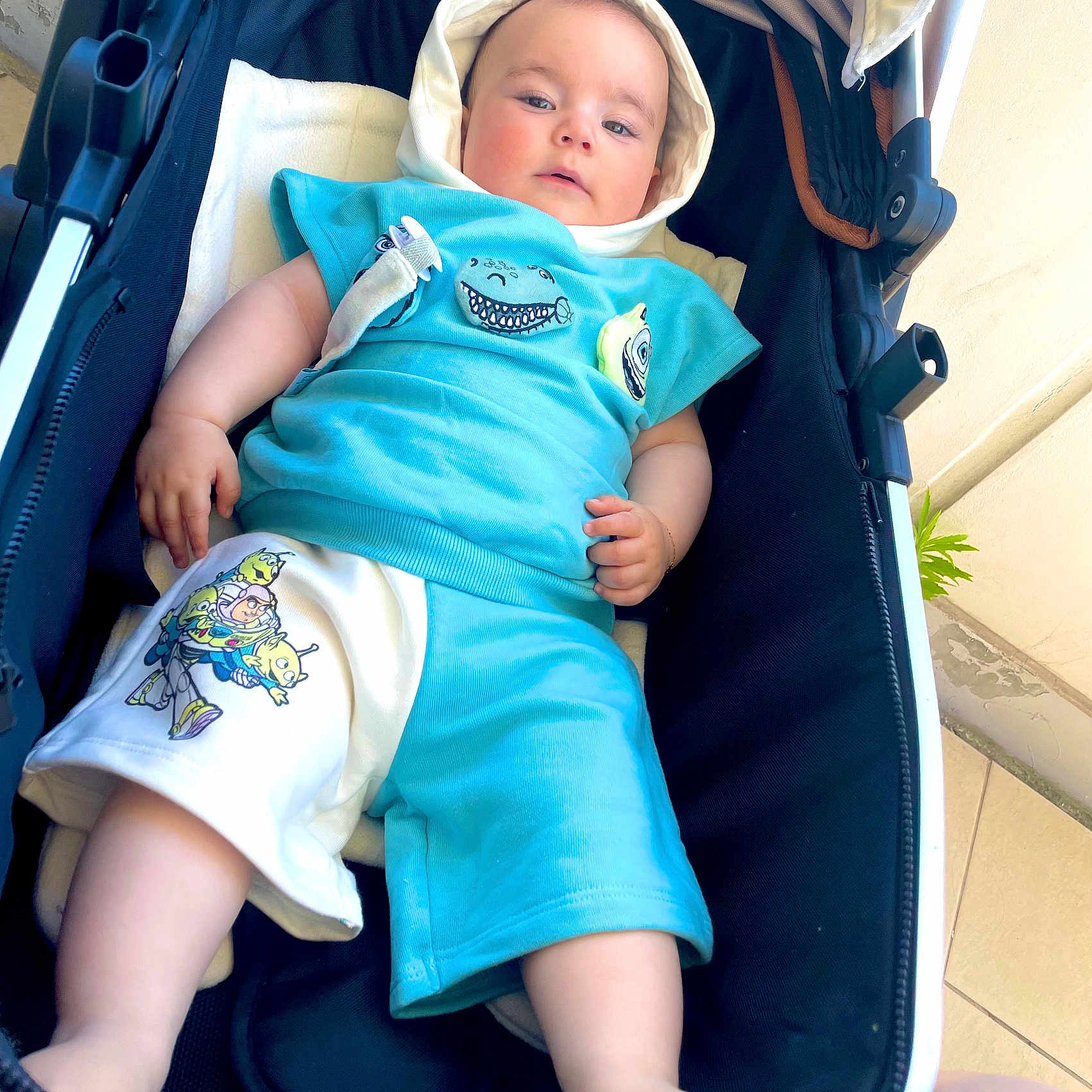 Valentino participe au concours pour gagner de l'argent avec cette photo : baby, blue_clothing, cartoon_print, child, cute, daylight, expression, face, footwear, hand, hood, infant, legs, outdoor, person, relaxed, seated, shorts, skin, stroller