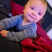 Riley joined the competition — help win amazing prizes! toddler, child, baby, blue_sweater, smile, face, hands, indoor, couch, cushion, red_blanket, portrait, young_child, cozy, curious, person, furniture, warm_lighting, closeup, happy