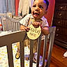 Amor is registered to the contest to win money with this photo: baby, bow_tie, child, clothing, crib, curtains, cute, dresser, floor, furniture, happy, holding, indoor, infant, person, room, smiling, standing, wooden_floor, wooden_sign