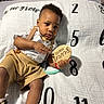 Amor is registered to the contest to win money with this photo: baby, blanket, brown_skin, child, clothing, curious, face, footwear, head, indoor, infant, milestone_blanket, numbers, person, short_hair, sitting, sneakers, suspenders, toy, wooden_sign