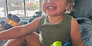 Asiah is registered to the contest to win money with this photo: toddler, child, smiling, indoor, bed, sippy_cup, toy_truck, curly_hair, window, quilt, clothing, happy, person, furniture, daylight, face, sitting, home, play, relaxed