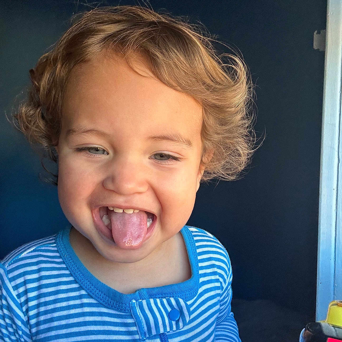 Asiah joined the competition — help win amazing prizes! background, blue_shirt, child, curly_hair, cute, expression, face, hands, happy, indoor, light, person, playful, portrait, skin, smiling, striped_clothing, toddler, tongue_out, young_child