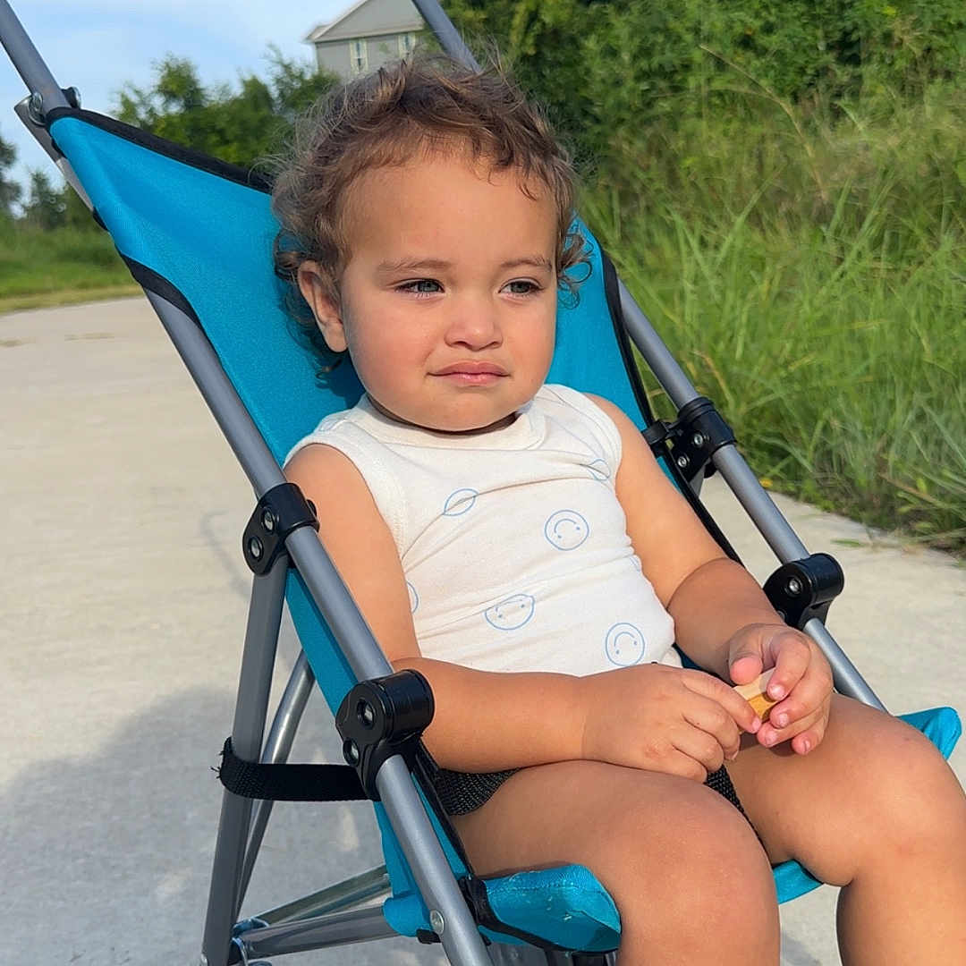 Asiah joined the competition — help win amazing prizes! baby, blue, casual_clothing, child, curly_hair, daytime, expression, greenery, nature, outdoor, person, relaxing, sidewalk, sitting, small_object, stroller, summer, sunlight, thoughtful, wheels