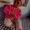 toddler, child, pink_sweater, beanie, curly_hair, indoor, checkered_floor, pink_shoes, casual_clothing, window, daylight, sitting, cute, young_child, portrait, floor_pattern, hands_clasped, expression, cozy, fashion