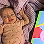 baby, cartoon, child, childrens_toy, close_up, face, floral_pattern, hand, happy, indoors, infant, lying_down, pacifier, pink_onesie, portrait, purple_blanket, skin, smiling, tablet, technology