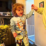 Harriet is registered to the contest to win money with this photo: toddler, child, curly_hair, bluey_onesie, indoor, table, hand_holding, barefoot, television, flower_vase, yellow_flowers, photograph, doorway, baby_gate, carpet, smiling, happy, person, furniture, family_moment