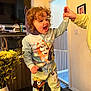 Harriet is registered to the contest to win money with this photo: toddler, child, curly_hair, onesie, indoor, table, holding_hand, smiling, television, vase, flowers, baby_gate, carpet, doorway, adult_hand, living_room, happy, standing, cartoon_clothing, home