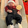 baby, infant, sleeping, santa_hat, plaid_shirt, suspenders, denim_pants, white_booties, wooden_sign, christmas_tree, holiday, festive, cushioned_chair, carpet, indoors, peaceful, cute, child, holiday_decor, seasonal