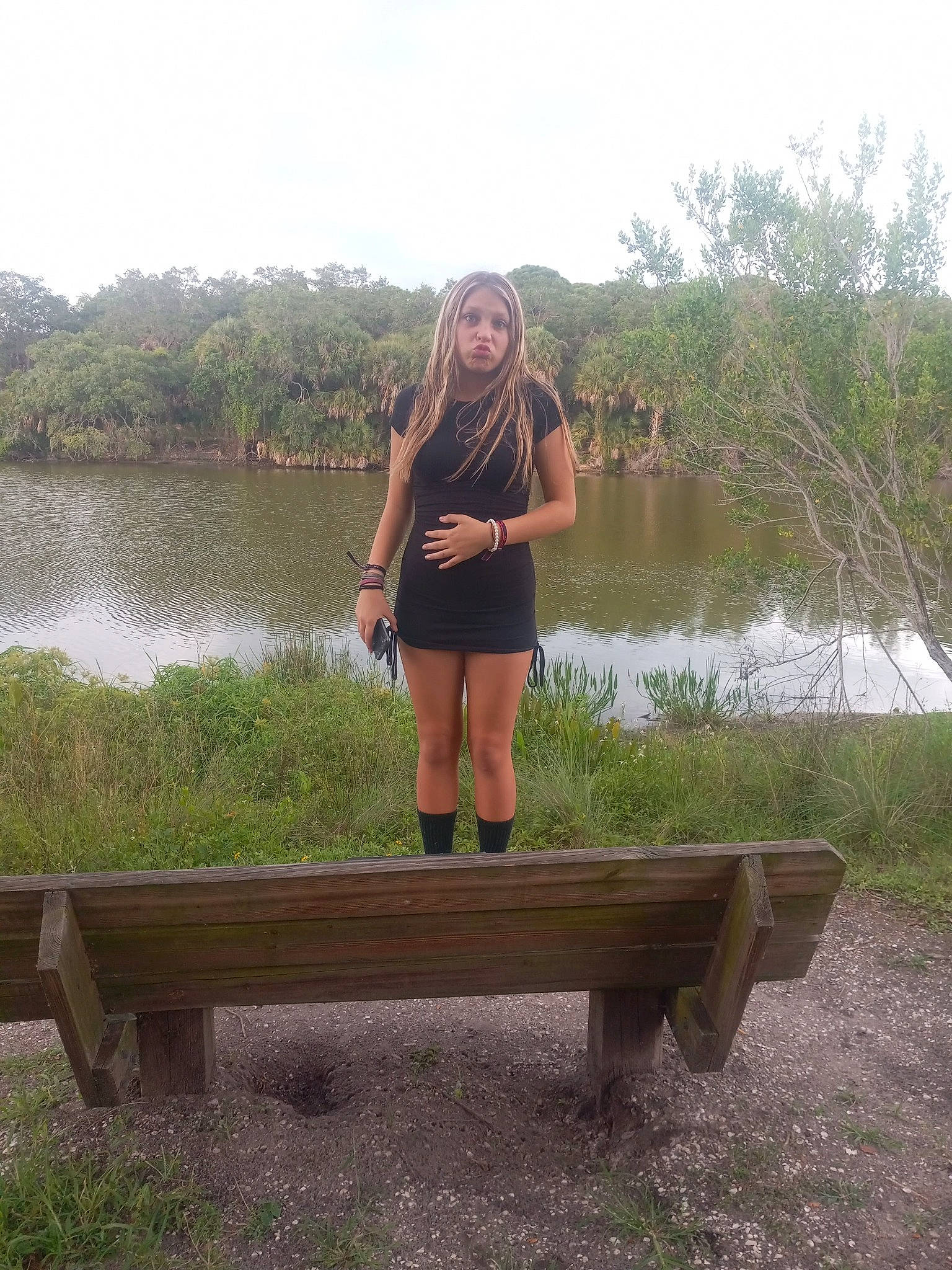 Rihanna is registered to the contest to win money with this photo: bank, bench, grass, grass_family, grassland, lake, landscape, leg, leisure, natural_landscape, outdoor_bench, outdoor_furniture, people_in_nature, person, plant, shoe, sky, thigh, tree, water