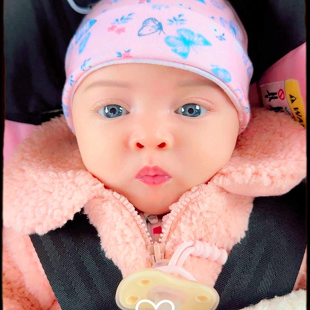 Kaiiv is registered to the contest to win money with this photo: baby, blanket, blue_eyes, car_seat, child, closeup, cute, expression, face, hat, indoors, infant, jacket, pacifier, person, pink_clothing, portrait, seatbelt, straps, winter_clothing