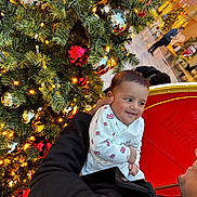 Rudvik is registered to the contest to win money with this photo: baby, child, christmas_tree, ornaments, red_ball, silver_ball, lights, holiday, smile, happy, person, indoor, festive, clothing, coat, hand, face, celebration, decoration, mall