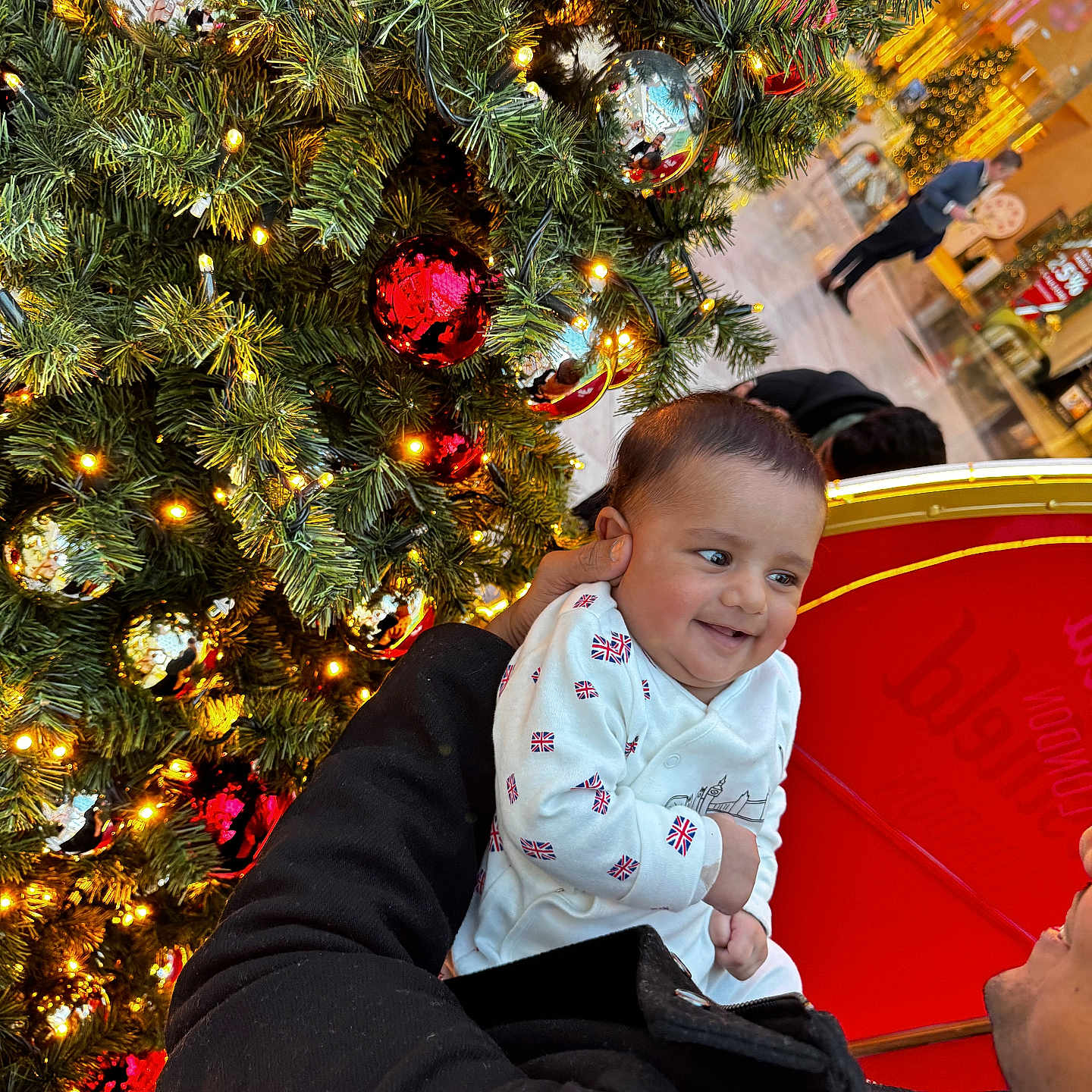 Rudvik is registered to the contest to win money with this photo: baby, celebration, child, christmas_tree, clothing, coat, decoration, face, festive, hand, happy, holiday, indoor, lights, mall, ornaments, person, red_ball, silver_ball, smile