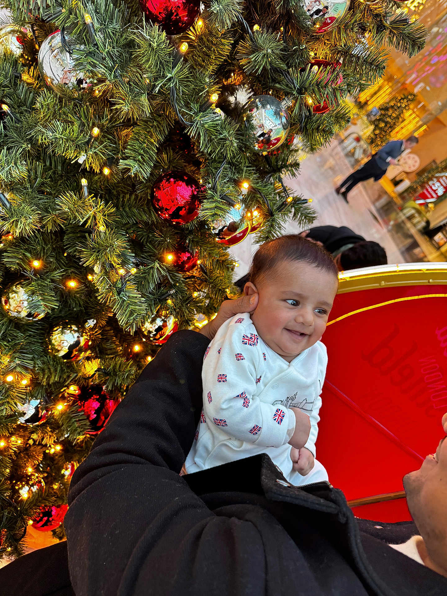 Rudvik is registered to the contest to win money with this photo: baby, child, christmas_tree, ornaments, red_ball, silver_ball, lights, holiday, smile, happy, person, indoor, festive, clothing, coat, hand, face, celebration, decoration, mall