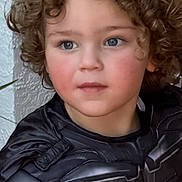 Nicholas is registered to the contest to win money with this photo: child, curly_hair, costume, batman, superhero, black_outfit, portrait, blue_eyes, hand, wall, texture, young_child, serious_expression, indoor, closeup, face, person, skin, cute, clothing