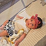 baby, infant, playpen, toy, giraffe, bow, headband, blanket, book, clothing, soft_toy, indoor, child, cute, lying_down, person, smiling, toddler, mesh, fabric