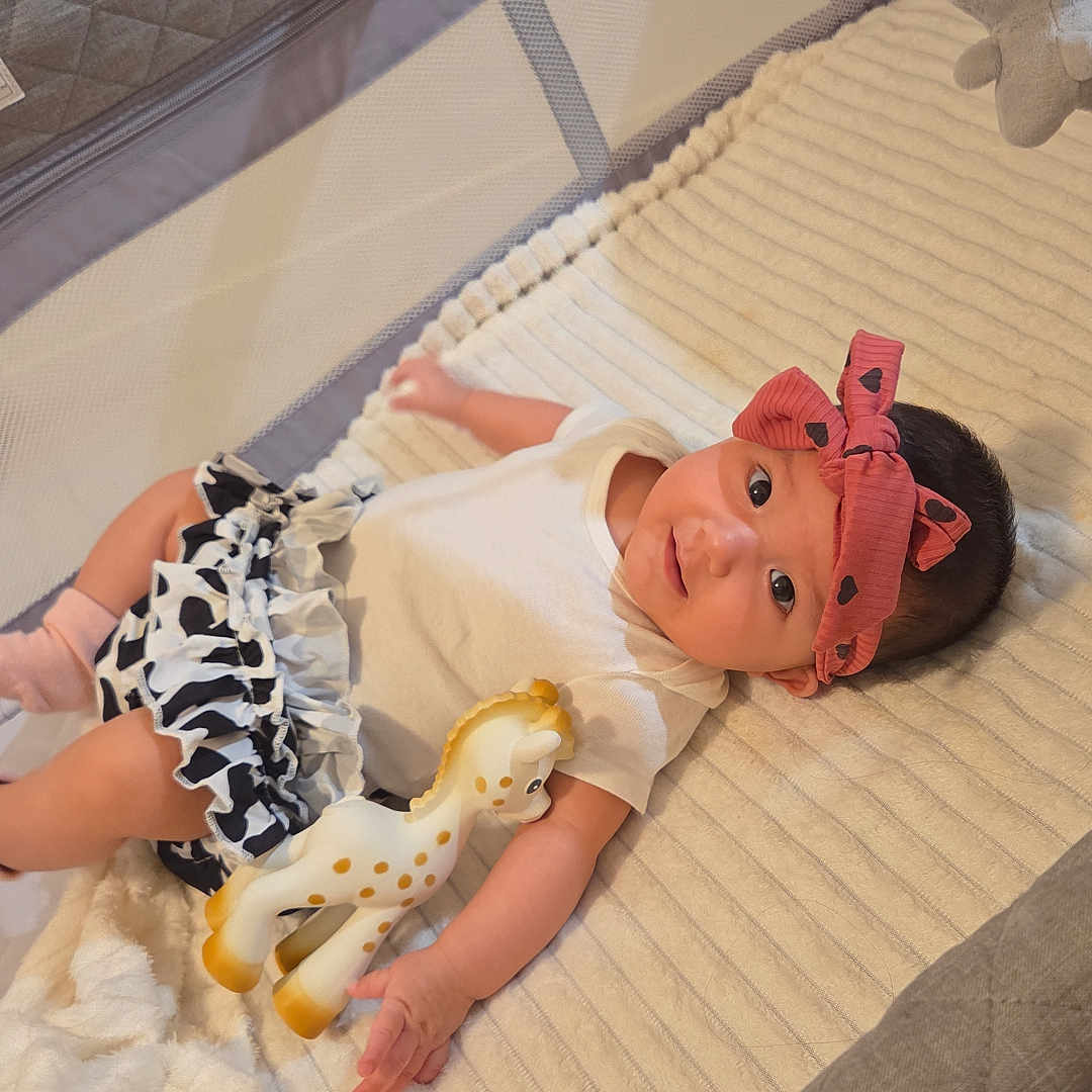 Sophia joined the competition — help win amazing prizes! baby, infant, playpen, toy, giraffe, bow, headband, blanket, book, clothing, soft_toy, indoor, child, cute, lying_down, person, smiling, toddler, mesh, fabric