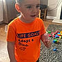 child, boy, tshirt, orange, indoor, floor, rug, toys, building_blocks, ice_cream, popsicle, shorts, socks, furniture, curious, standing, hair, face, light, expression