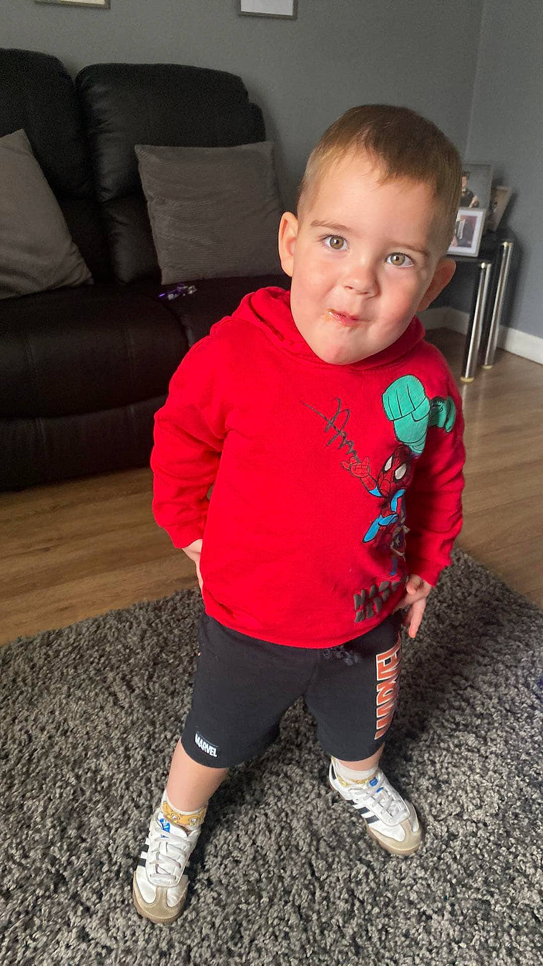 Mason is registered to the contest to win money with this photo: boy, carpet, casual_clothing, child, couch, floor, home, indoor, living_room, marvel, person, photography, pillow, playful, pose, red_hoodie, shorts, smile, sneakers, wooden_floor