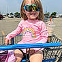 child, shopping_cart, sunglasses, pink_shirt, rainbow_design, parking_lot, drink, sunny, smile, happy, outdoor, reflection, casual_clothing, legs, hand, daytime, retail, store_parking, person, summer