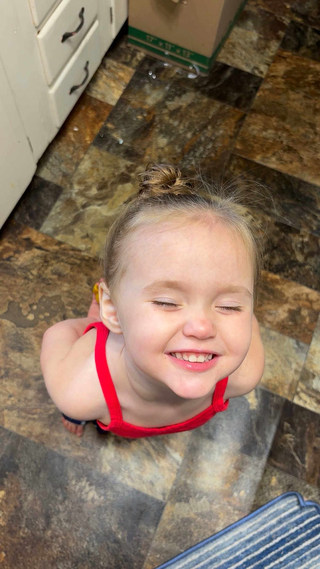 Rae is registered to the contest to win money with this photo: child, smiling, happy, closed_eyes, red_clothing, standing, kitchen, floor_tiles, blond_hair, hair_bun, indoor, cute, person, young_child, playful, casual, small_child, joy, portrait, top_view