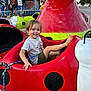 child, toddler, smiling, amusement_park, ride, ladybug, red, green, shorts, tshirt, happy, outdoor, daylight, people, fence, superhero_mural, background, fun, playful, leisure