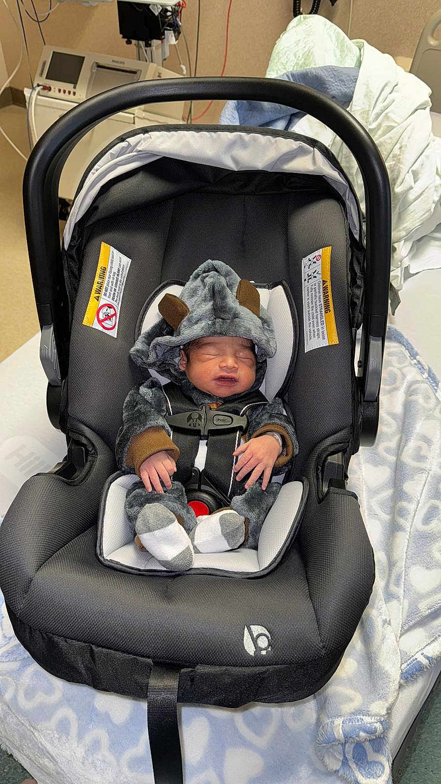 Levy is registered to the contest to win money with this photo: baby, infant, car_seat, onesie, sleeping, blanket, hospital, medical_equipment, newborn, clothing, soft_toys, cute, person, bed, fabric, headwear, indoors, resting, child, seatbelt